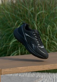 Μαύρο αθλητικό παπούτσι με λείο συνθετικό επάνω μέρος, υφασμένο μεσαίο μέρος, αντανακλαστικές λεπτομέρειες και μαλακό περιλαίμιο. Εμφανίζεται στον αέρα πάνω από ξύλινη επιφάνεια.