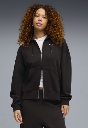 Vrouw met krullend haar, gekleed in een zwart vest met ritssluiting en zwarte broek, staand tegen een effen grijze achtergrond.