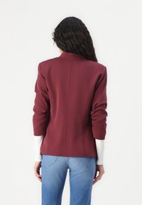 Blazer bordeaux avec épaules structurées, manches retroussées et coupe ajustée. Associé à un jean bleu clair, présentant un design simple et épuré.