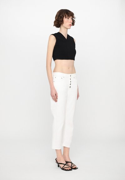 Top corto nero con orlo a scallop, abbinato a jeans bianchi a gamba larga con chiusura a bottoni. Il modello indossa tacchi neri con cinturini.