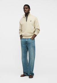 Άνδρας που φοράει μπεζ φούτερ με φερμουάρ μέχρι το στήθος και λογότυπο ζώου, μπλε τζιν, καφέ σανδάλια και γυαλιά, στέκεται με τα χέρια στις τσέπες.