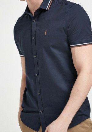 Man wearing short-sleeve navy button-up shirt with white striped collar and sleeve edges, and small embroidered logo on chest.