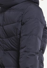 Mørkeblå puffy jakke med hætte, med vandret quiltede sektioner, en lynlåslomme på siden, og en glat, shiny overflade.