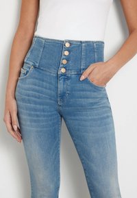 Jeans de mezclilla azul de cintura alta con cuatro botones dorados, bolsillos frontales y una textura desgastada en la tela.