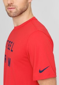 Hombre vistiendo una camiseta roja de Nike con texto en azul marino parcialmente visible y un logo de swoosh de Nike en azul marino en la manga, contra un fondo gris.