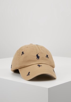 Casquette de baseball beige avec motifs de crânes et os croisés noirs, ainsi qu'un logo de joueur de polo brodé en noir à l'avant, posée sur une surface blanche.