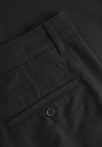 Black trousers featuring a rear pocket with a button closure and an embossed logo. Fabric exhibits a smooth, textured finish.