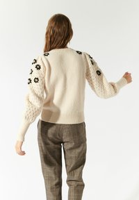 Maglione a maglia di un beige chiaro con maniche a sbuffo testurizzate e ricami floreali neri. Orlo e polsini a costine. Indossato con pantaloni a quadri grigi.