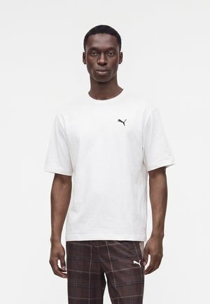 Άνδρας που φοράει λευκό t-shirt Puma και καφέ καρό παντελόνι, στέκεται μπροστά σε απλό λευκό φόντο.