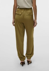 Pantalons verts olive, ajustés avec une taille élastique, deux poches arrière et des poignets froncés, associés à des chaussures à talons bruns.