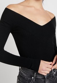 Haut noir côtelé à épaules dénudées avec manches longues, présentant un décolleté en V profond et un tissu texturé. Porté avec un jean taille haute.