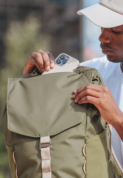 Man som bär vit keps lägger sin smartphone i framfickan på en olivgrön ryggsäck utomhus.