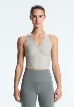 Kobieta w beżowym, bezrękawnym topie z dekoltem w serek i szarych legginsach z wysokim stanem, stojąca na tle jasnego, jednolitego tła, zwrócona przodem.