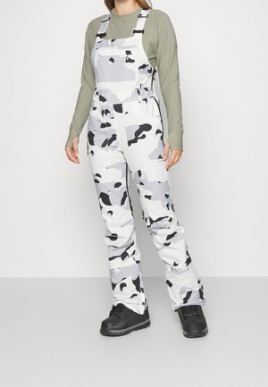 Person iført hvidt og gråt camouflage-buksesele, olivengrøn langærmet skjorte og sorte støvler, stående foran en ensfarvet baggrund.