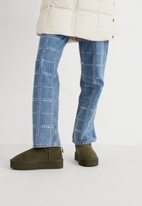 Olivno zeleni semiš škornji s zaobljenim nosom, ravnim podplatom in božnim zadrgo, oblečeni s svetloplavimi jeans hlačami z zatiči v mrežastem vzorcu.