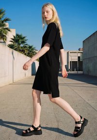 Blonde woman walking outdoors in a black knee-length dress and black platform sandals, with modern concrete buildings and palm trees nearby.