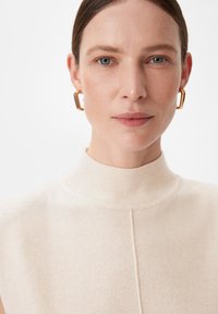 Beżowy top bez rękawów z wysokim kołnierzem, o prążkowanej fakturze i centralnym szwie; złote geometryczne kolczyki dopełniają stylizację.