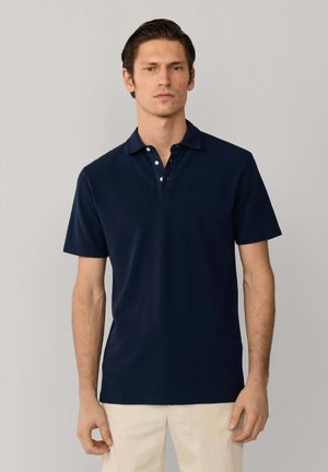 Hombre de cabello oscuro que lleva una camisa tipo polo de manga corta azul marino y pantalones beige, de pie contra un fondo claro y liso.