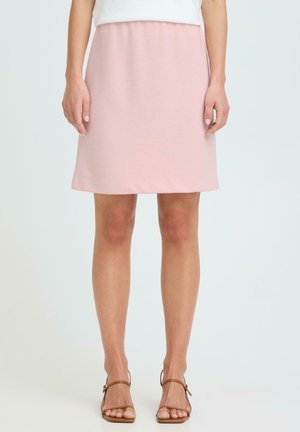 Mujer con falda rosa claro hasta la rodilla, blusa blanca y sandalias marrones de tacón abierto, sobre un fondo liso.