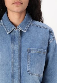 Blå denimjakke med spiss krage, vertikal knappelukking og brystlomme. Har subtil fading og kontrasterende søm.