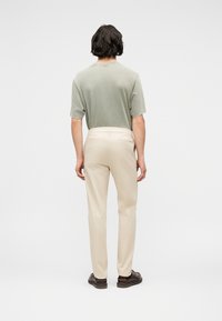 Homme aux cheveux noirs portant une chemise à manches courtes vert clair, un pantalon beige et des chaussures marron foncé, debout de dos sur un fond uni.