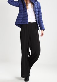 Prošívaná modrá bunda s vysokým límcem, černé kalhoty s širokými nohavicemi a pod ní bílá košile. Texturovaná látka a elegantní silueta.