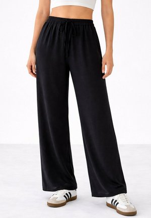 Personne portant un pantalon noir à jambes larges avec une taille élastique et des baskets blanches avec des rayures noires, debout sur un sol clair.