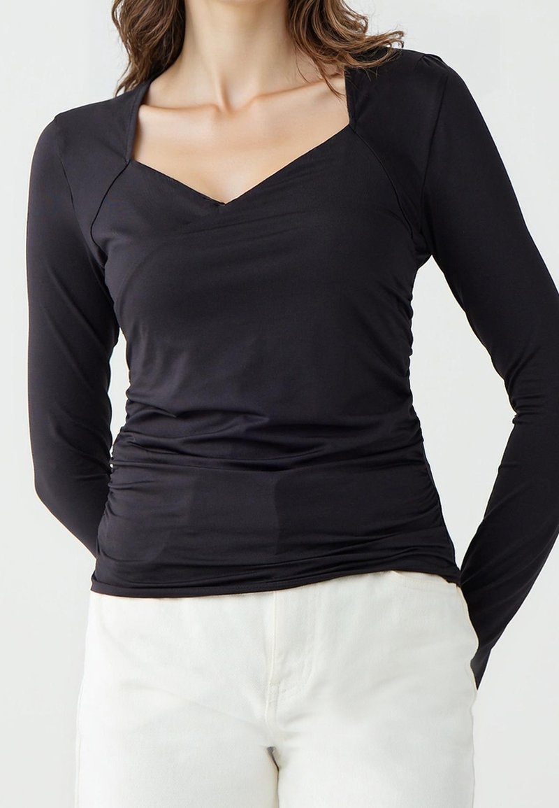 Femme portant un haut noir ajusté à manches longues avec un col carré et un pantalon blanc sur un fond uni.