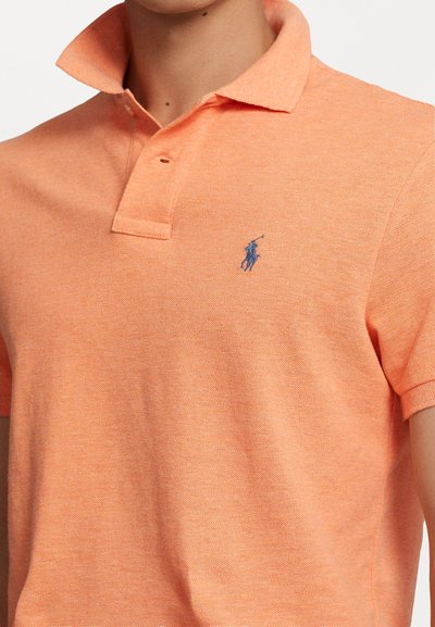 Tricou polo de culoare portocalie deschis, realizat dintr-un material texturat. Are un guler clasic, o bandă cu două butoane și un mic logo albastru pe piept.