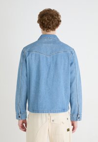 Jasnoniebieska kurtka dżinsowa o klasycznym kroju, z charakterystycznym kołnierzykiem, długimi rękawami i detalami szycia na plecach oraz ramionach.