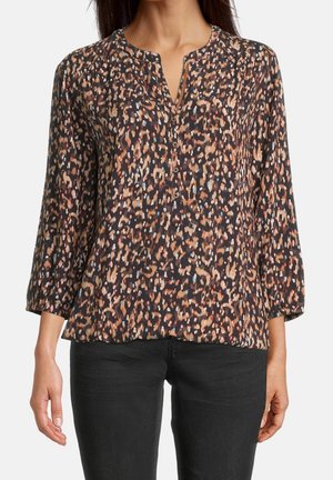 Vrouw draagt een blouse met lange mouwen en knopen met luipaardprint en een zwarte broek tegen een effen achtergrond.