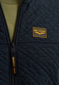 Marineblaue, gesteppte Zip-Jacke mit Rundhalsausschnitt, olivgrünem Innenfutter und gelbem PME Legend-Logo-Patch auf der Brust für Markenidentität.