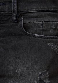 Jeans de mezclilla negra con una textura desgastada, con un bolsillo frontal y detalles de costura. La tela está ligeramente descolorida en algunas áreas.