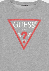 Camiseta de algodón gris con un logo triangular rojo que incluye el texto blanco "GUESS" y "U.S.A." sobre un signo de interrogación rojo, con números debajo.