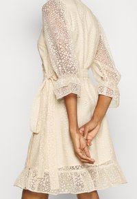 Robe de couleur crème avec manches bouffantes en mousseline, tissu texturé, taille cintrée par une ceinture, et ourlet volanté. Le motif présente des détails en pointillés doux.