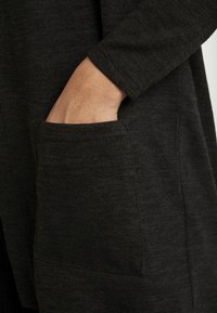 Prenda texturizada de color gris oscuro con un bolsillo lateral, de corte ajustado y con un detalle de puño redondeado en la manga larga.