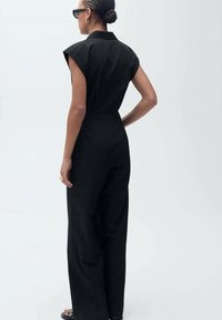 Svart skräddarsydd jumpsuit med korta ärmar, åtsittande midja och vida byxor. Tyget verkar slätt med subtil textur och sömmar.