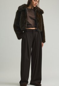 Giacca corta in pelliccia sintetica marrone con chiusura a zip, abbinata a pantaloni marroni a vita alta e gamba larga. Texture liscia in tutta la superficie.