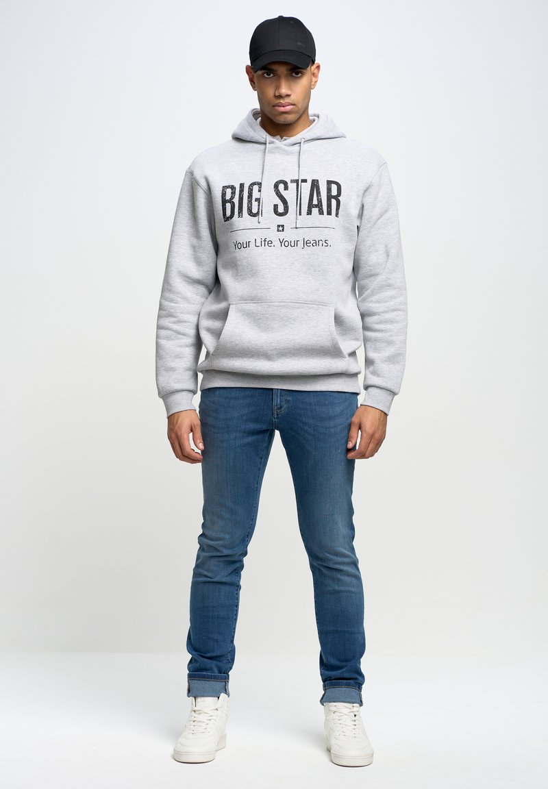 Šedá mikina s kapucí a nápisem "BIG STAR", přední kapsou a stahovací šňůrkou; spárována s modrými džínami a bílými teniskami; model má na hlavě černou kšiltovku.