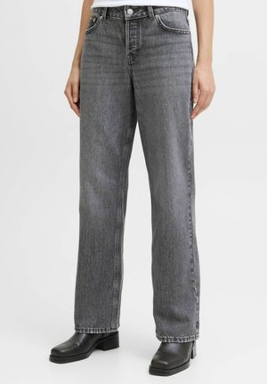 Femeie purtând blugi gri cu talie înaltă și croială dreaptă, asortați cu botine negre cu toc, stând în fața unui fundal simplu.