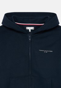 Mornaric blue zip-up dukserica s kapuljačom, prednjim džepovima i malim logom "Tommy Hilfiger EST. 1985" na prsima.