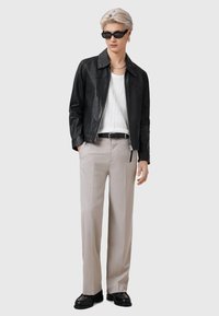 Zwarte leren jas, witte ribbed tanktop, lichtbeige broek, zwarte riem, zwarte instappers, draagt donkere zonnebril. Minimalistische stijl.