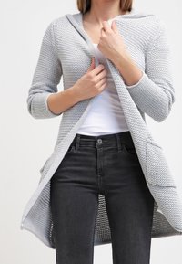 Světlounce šedý pletený cardigan s kapucí, otevřený vpředu, s dlouhými rukávy a bočními kapsami, nošený přes bílý top a tmavé džíny.