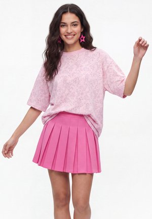 Ung kvinde med langt mørkt hår smiler, iført en lyserød mønstret T-shirt, hot pink plisseret nederdel og stjerneformede øreringe.