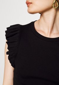 Top canotta nero a coste con dettagli arricciati sulle spalle. La scollatura è rotonda e il tessuto appare liscio con un design aderente.