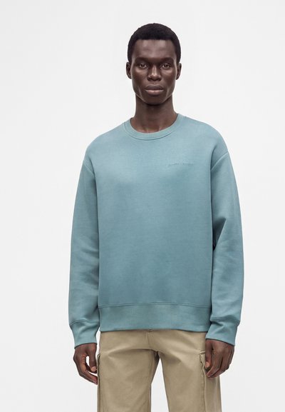 Jeune homme aux cheveux courts porte un sweat-shirt bleu clair et un pantalon cargo beige, debout devant un fond clair uni.