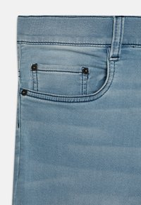 Jeans in denim azzurro chiaro con tasca anteriore, componenti metallici neri e cuciture a contrasto. La texture del tessuto è liscia con una leggera sfumatura.