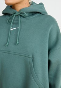 Πράσινη κουκούλα φτιαγμένη από μαλακό ύφασμα, με μπροστινή τσέπη, ρυθμιζόμενα κορδόνια και μια διακριτική λευκή λογότυπο Nike.