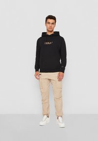 Sort hættejakke med snoretræk og logo, beige cargo-bukser med sidelommer og hvide sneakers. Glat stof, normal pasform.
