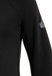 Parte superior de neopreno negra con un diseño ajustado. Presenta costuras planas y un acento de logo gris en el hombro. Material suave y elástico.